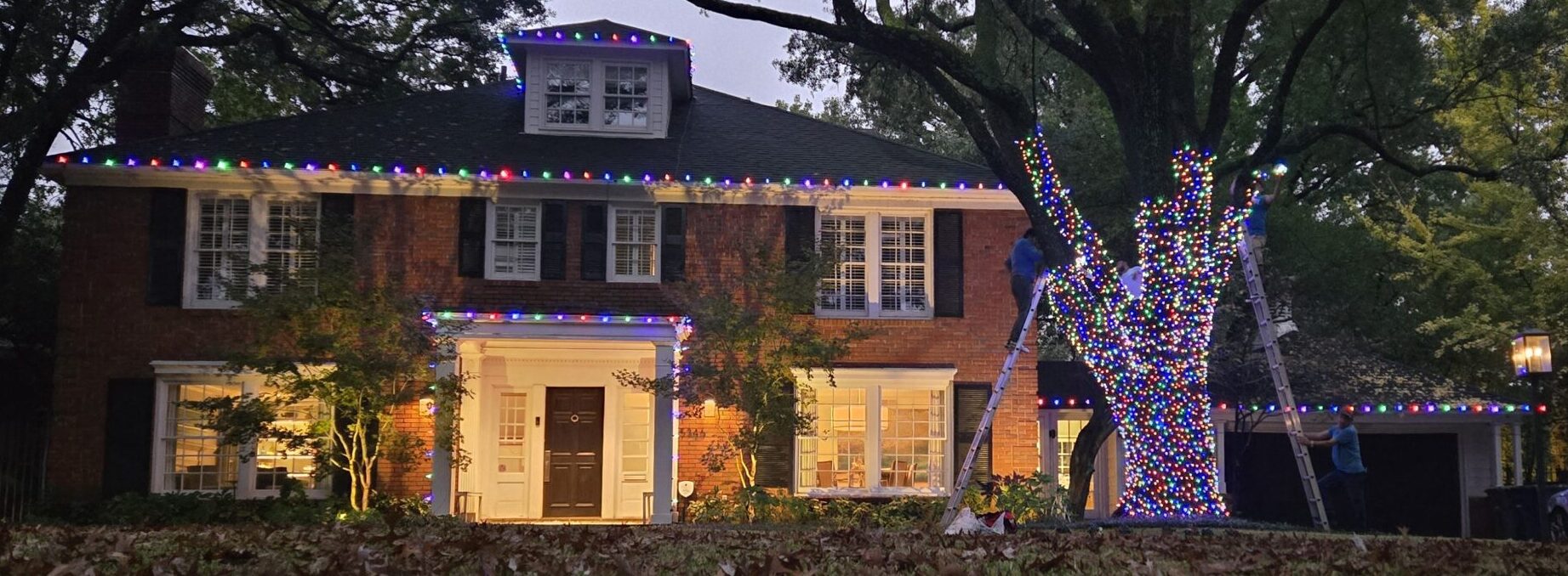 Brick two-story house with multicolored holiday lights along the roof and a tree wrapped in lights; workers on ladders decorate at dusk in a suburban yard.